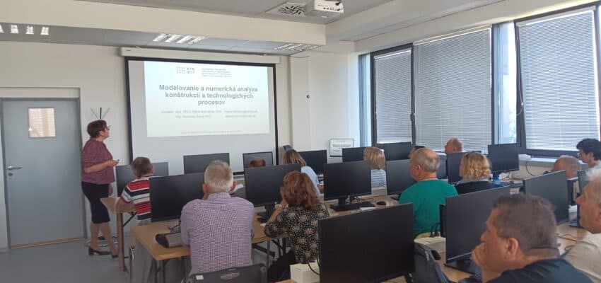 Na obrázku sú Vzdelávajúci sa učitelia sediaci za počítačmi v pozadí stojaca školiteľka pozerajúca sa na prezentáciu na tabuli