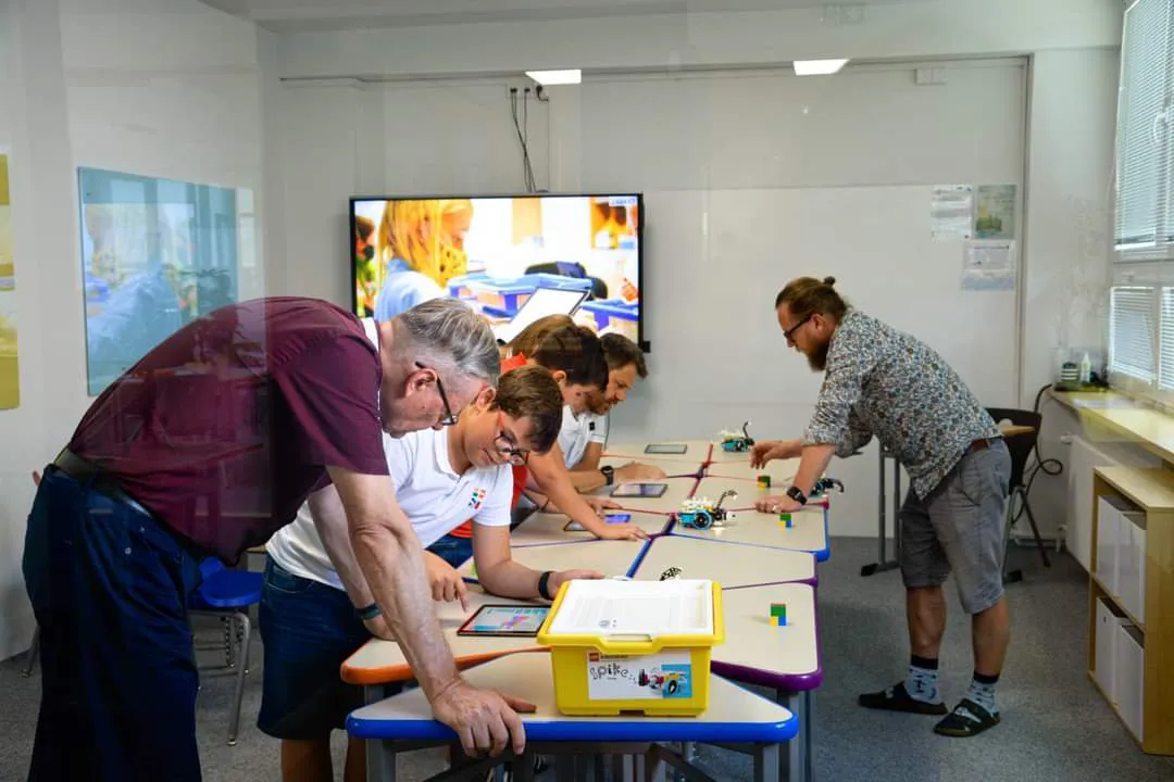 Na fotke viacero ľudí (žiakov/účastníkov) pracuje pri stoloch s tabletmi.