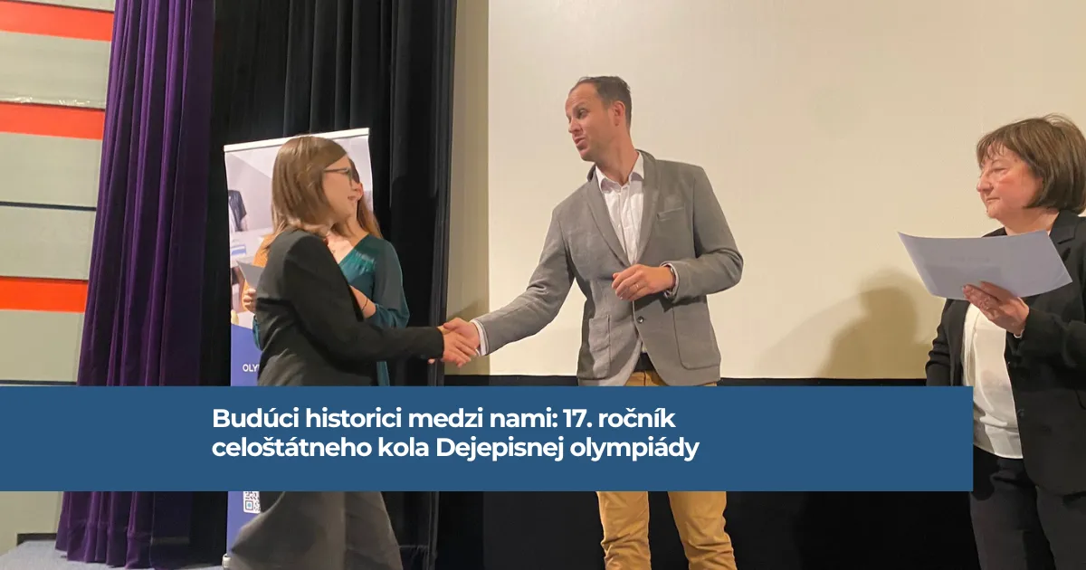 Na obrázku riaditeľ podáva ruku žiačke. Žena po jeho lavici drží v ruke diplom. V pozadí je reklamný banner a ďalšia žena