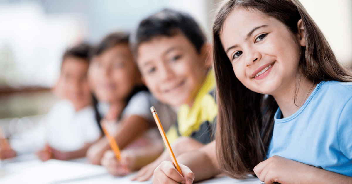 Na fotografii sú štyri deti, ktoré sa pozerajú do kamery a usmievajú. V ruke držia ceruzku a sedia za stolom.
