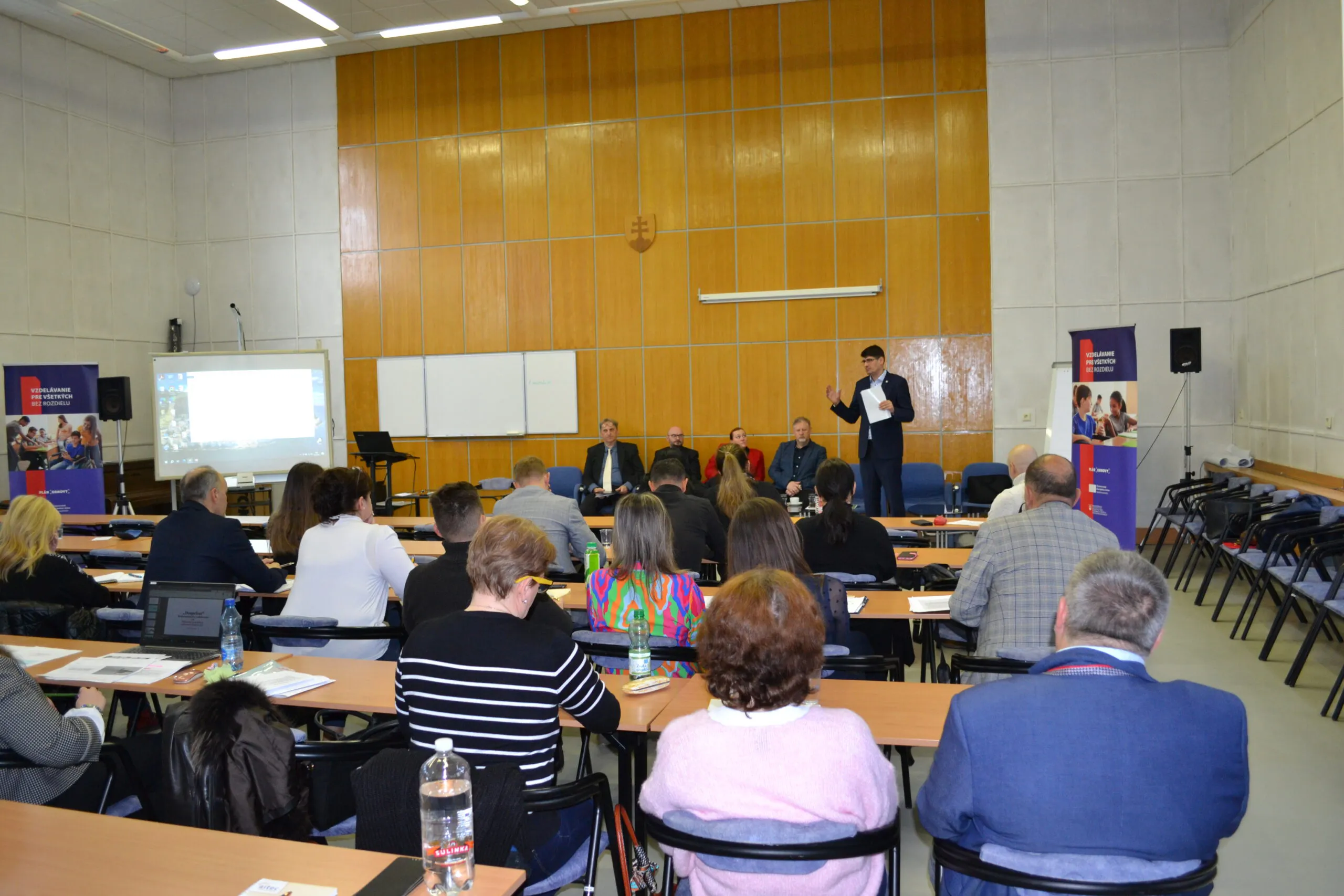 Ľudia sediaci v sále, pred nimi stojí rečník.