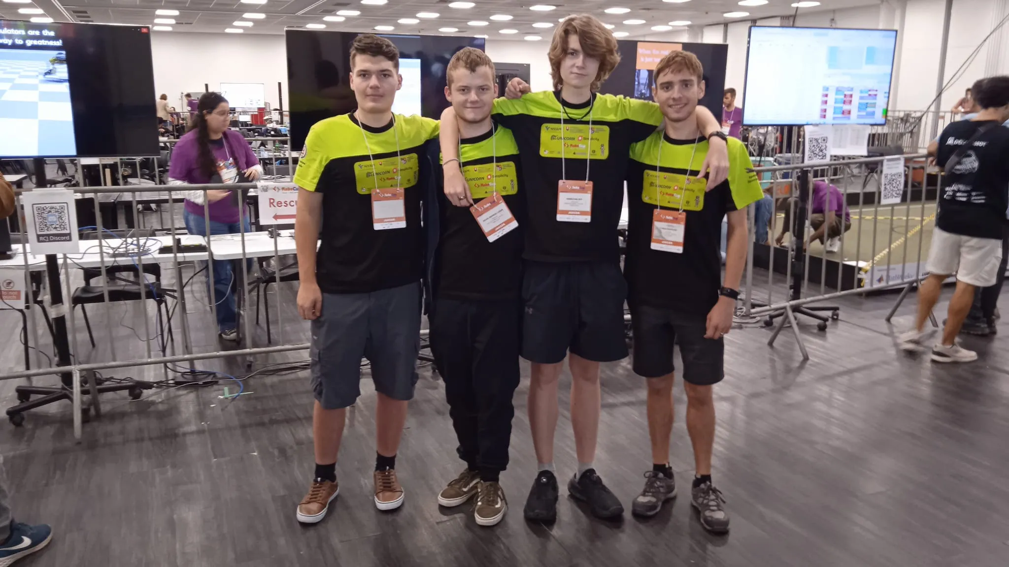 Na obrázku stoja štyria chlapci, držia sa okolo pliec a usmievajú sa do objektívu. V pozadí sú iní ľudia a veľké obrazovky.