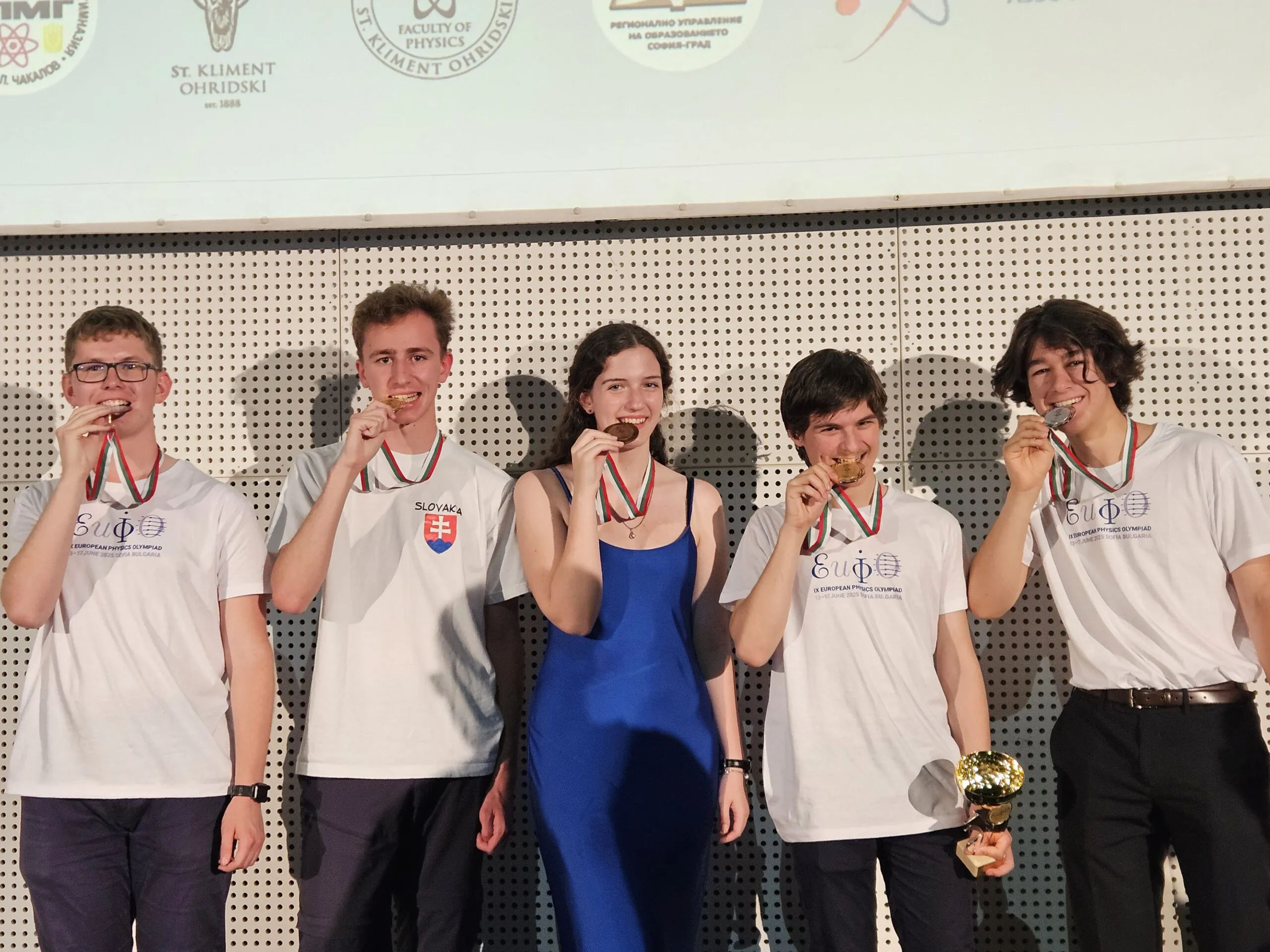 Na fotke sa nachádzajú štyria chlapi a jedno dievča, všetci držia pri ústach medaile zo súťaže a usmievajú sa.