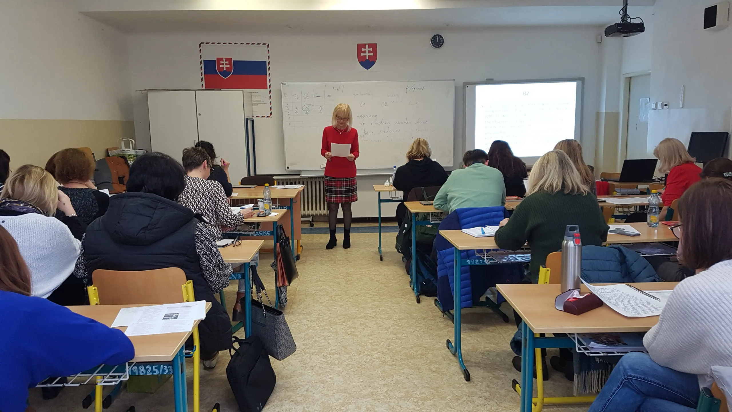 Žena číta z poznámok pred poslucháčmi, ktorí si zapisujú.