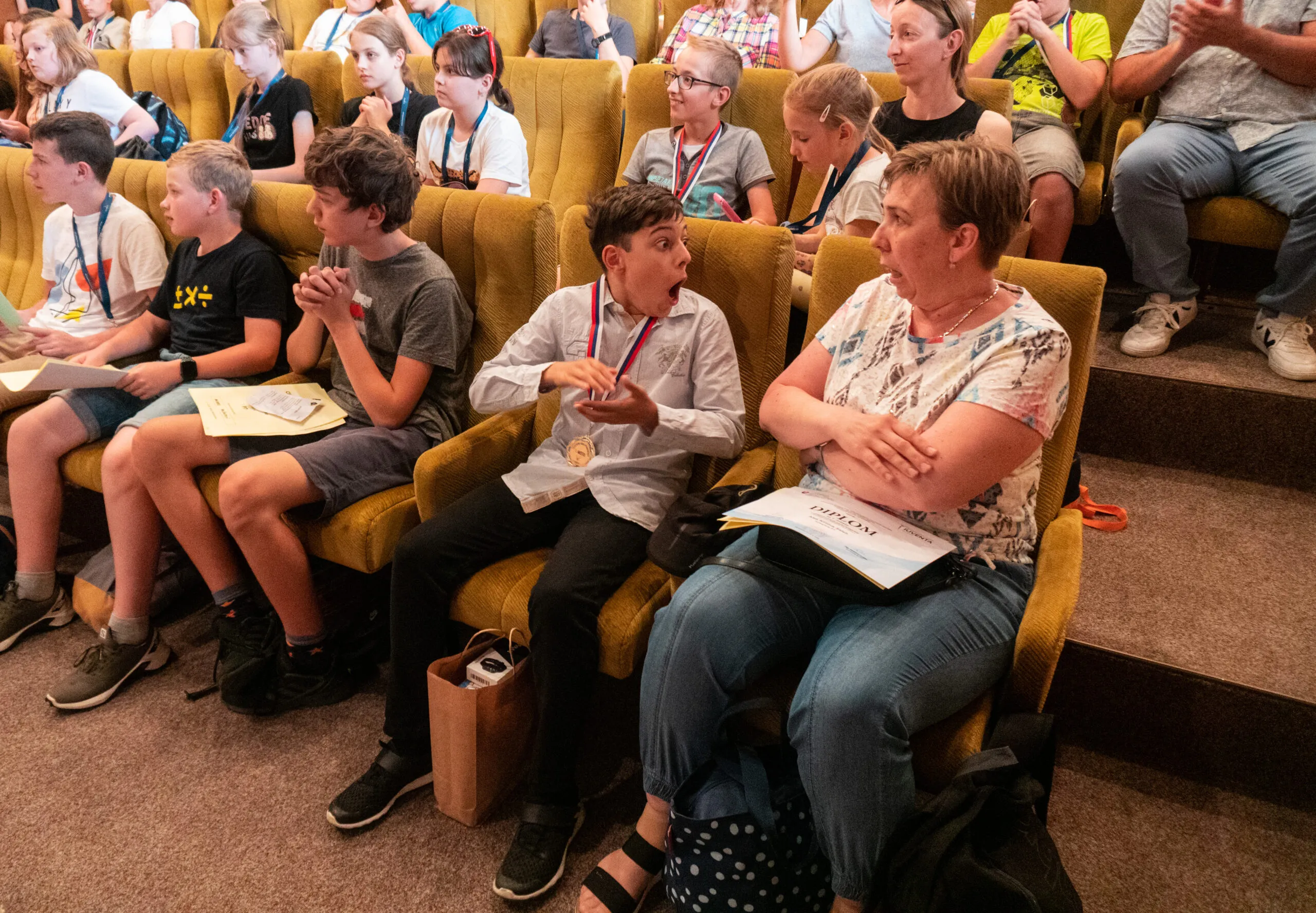 Zasadacia sála plná súťažiacich, ktorí sedia na stoličkách rad za radom a sledujú vyhodnotenie.