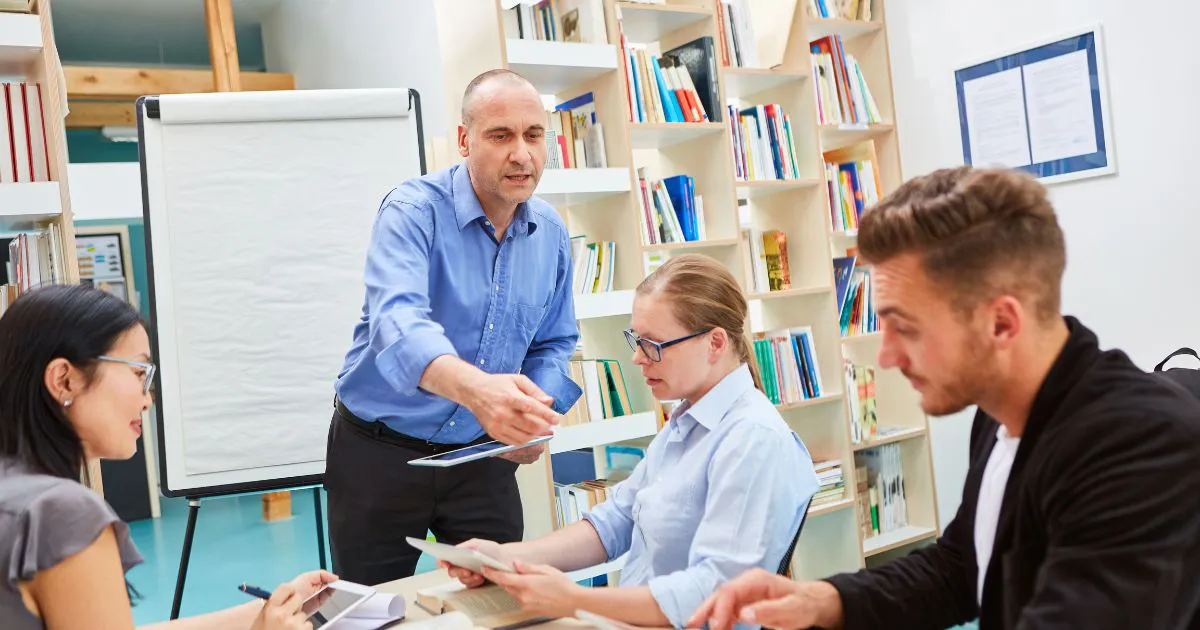 Ilustračný obrázok výučby, človek pred tabuľou vysvetľuje ostatným, ktorí ho počúvajú.