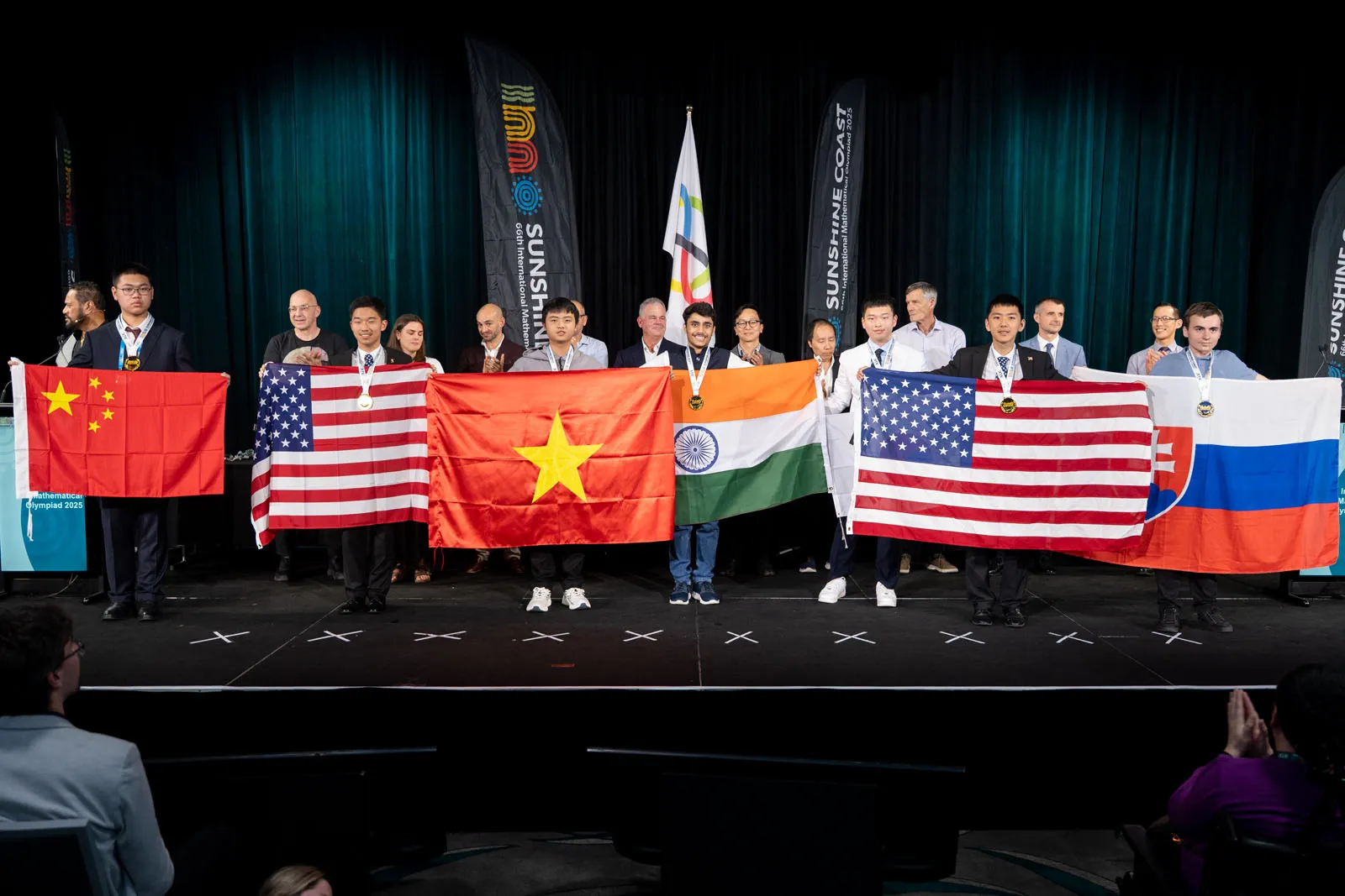 Na obrázku sa nachádza jedenásť osôb z toho päť chlapcov vpredu drží vlajky svojich krajín. Ľudia z publika si ich fotografujú.
