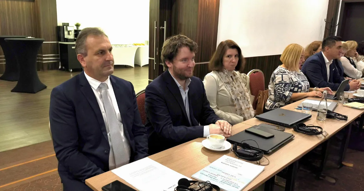 Na fotografii sedia ľudia za stolmi zoradenými vedľa seba. PRed sebou majú na stole položené notebooky a papiere a pozerajú sa pred seba.