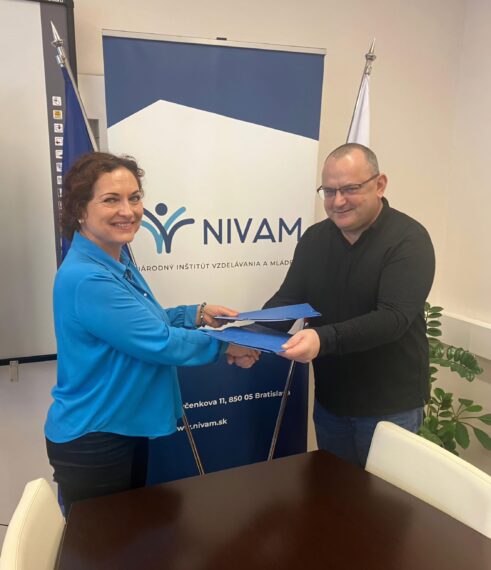 Na obrázku je PhDr. Romana Kánovská , generálna riaditeľka NIVaM a Ing. Peter Beck, poradca predsedu ZMOS, ktorí si podávajú ruky a zároveň si vymieňajú modré zakladače s dokumentmi, v pozadí sa nachádza banner s logom a názvom organizácie NIVaM.